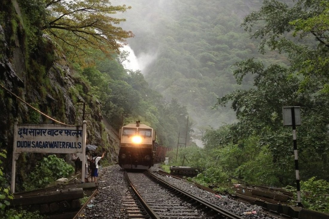 A train approaches Dudhsagar station through monsoon mist on the Braganza Ghats, with the bilingual station sign on the left