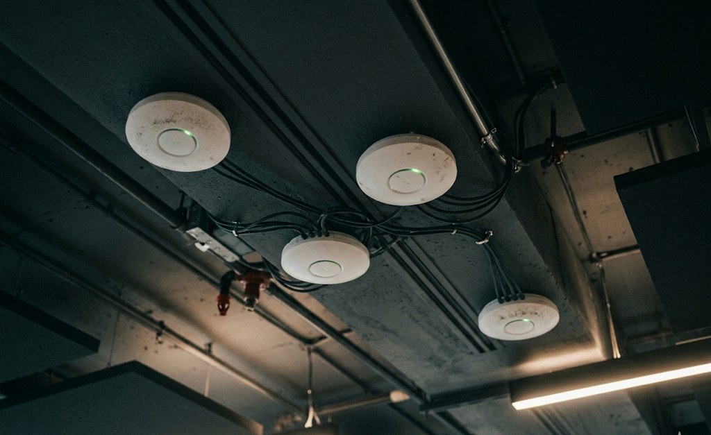 Four white Wi-Fi access points mounted close together on an exposed concrete ceiling, with cables looping between them and small green status LEDs glowing — visibly more APs in a small area than the space requires